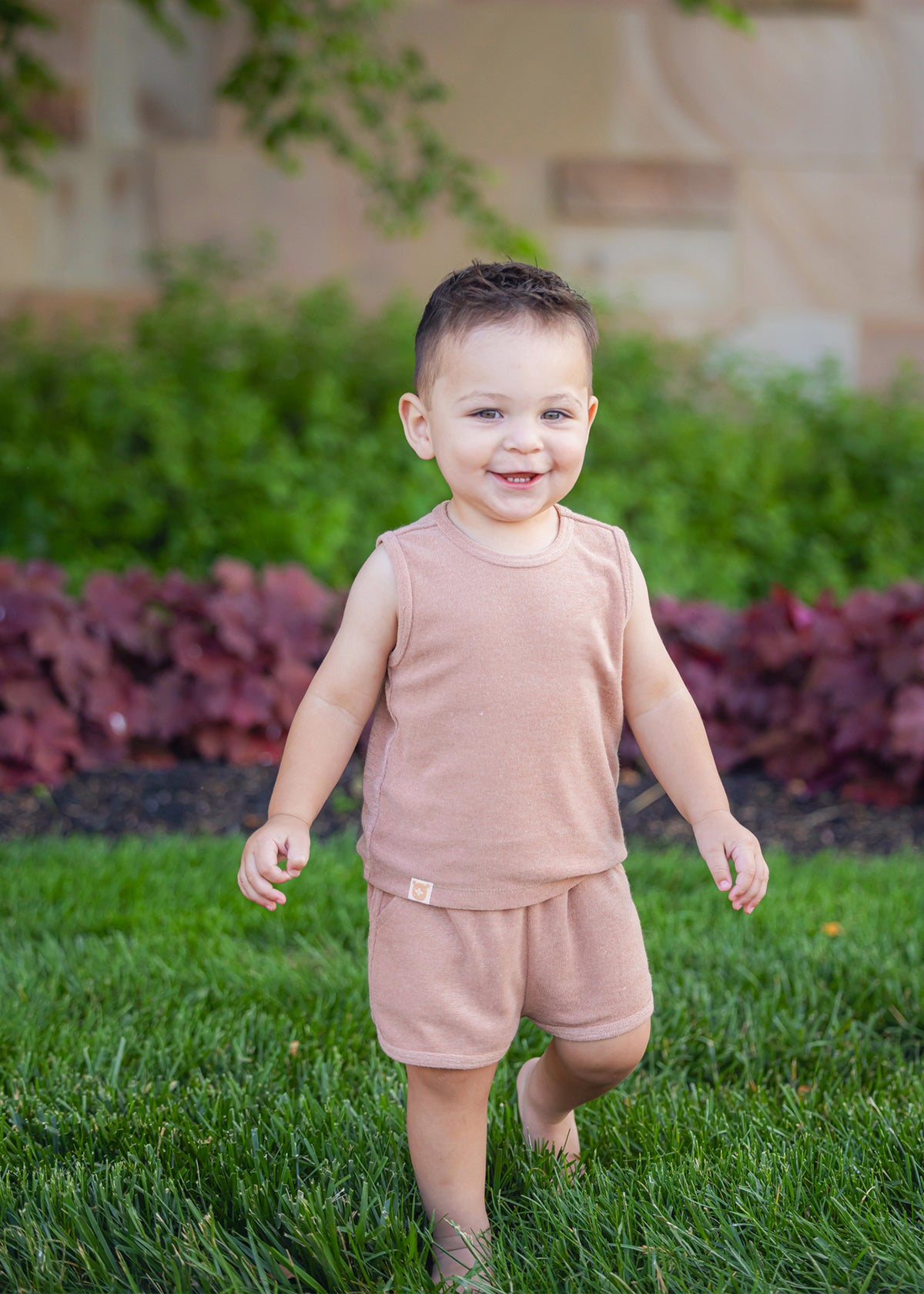 A smiling toddler stands barefoot on green grass, wearing the Beckett and Bear Dustin Sleeveless Two Piece Set. Bushes and a stone wall create a charming backdrop for this adorable kids’ outfit.