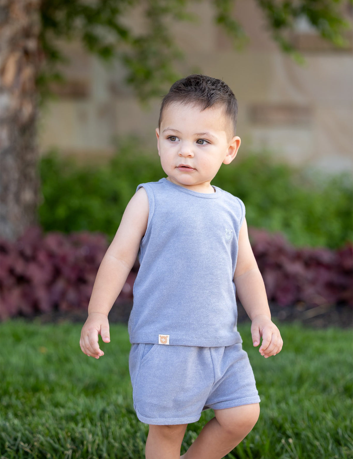 A young child wears the Axel Sleeveless Two Piece Set by Beckett and Bear, standing on grass outdoors with green plants, a tree, and blurred landscaping in the background.