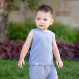 A young child wears the Axel Sleeveless Two Piece Set by Beckett and Bear, standing on grass outdoors with green plants, a tree, and blurred landscaping in the background.