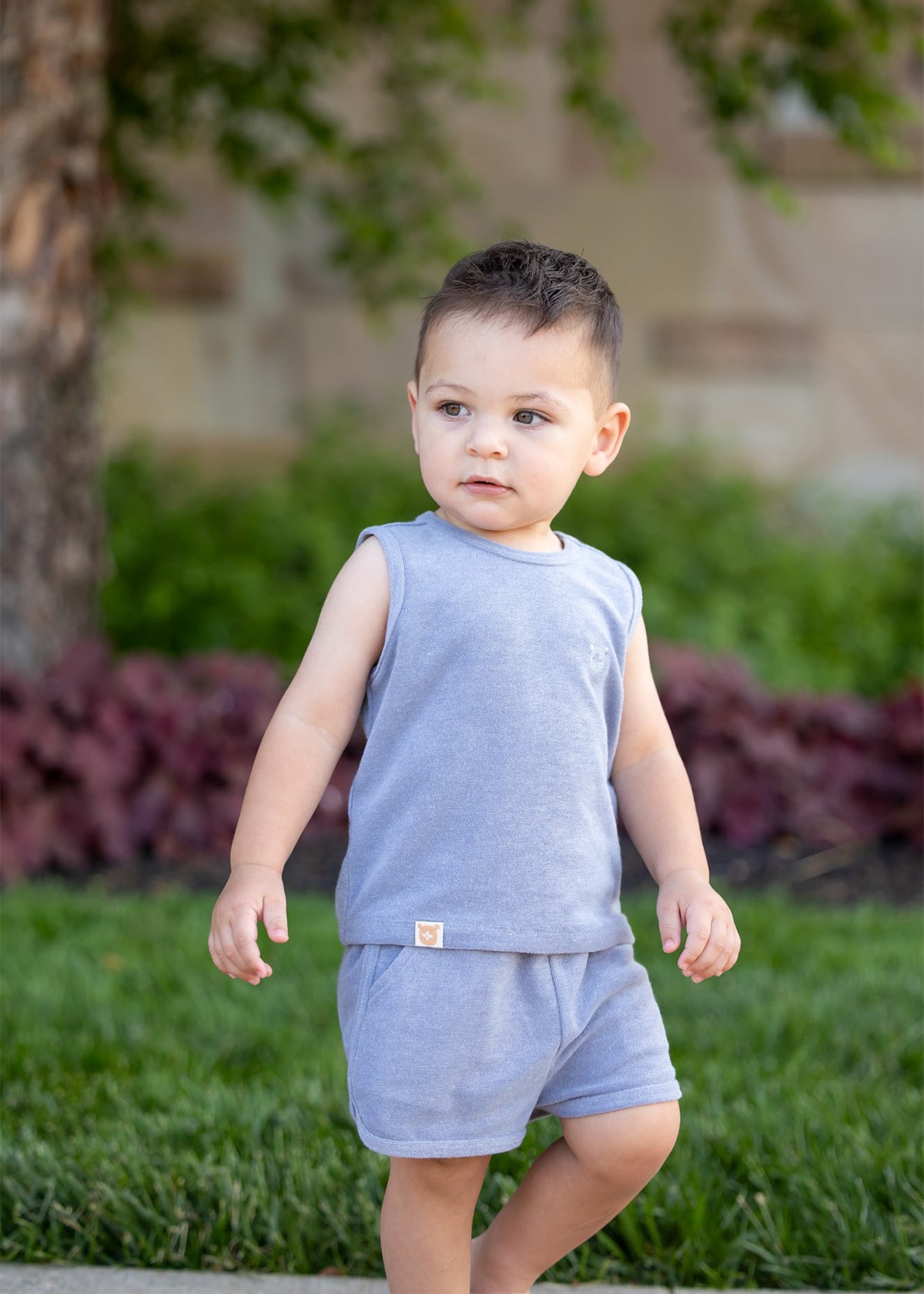 A young child wears the Axel Sleeveless Two Piece Set by Beckett and Bear, standing on grass outdoors with green plants, a tree, and blurred landscaping in the background.