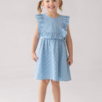 A smiling young girl with light brown hair in pigtails wears the Mabel and Honey Darling Denim Sleeveless Dress—a charming light blue dress with a star pattern and ruffled sleeves—paired with white sandals, standing against a plain light background.