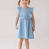 A smiling young girl with light brown hair in pigtails wears the Mabel and Honey Darling Denim Sleeveless Dress—a charming light blue dress with a star pattern and ruffled sleeves—paired with white sandals, standing against a plain light background.