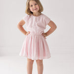 A young girl with short blonde hair smiles, hands on hips, wearing the Mabel and Honey Girlpop Dropshoulder Two Piece Set Pink and sparkly shoes against a plain white background.