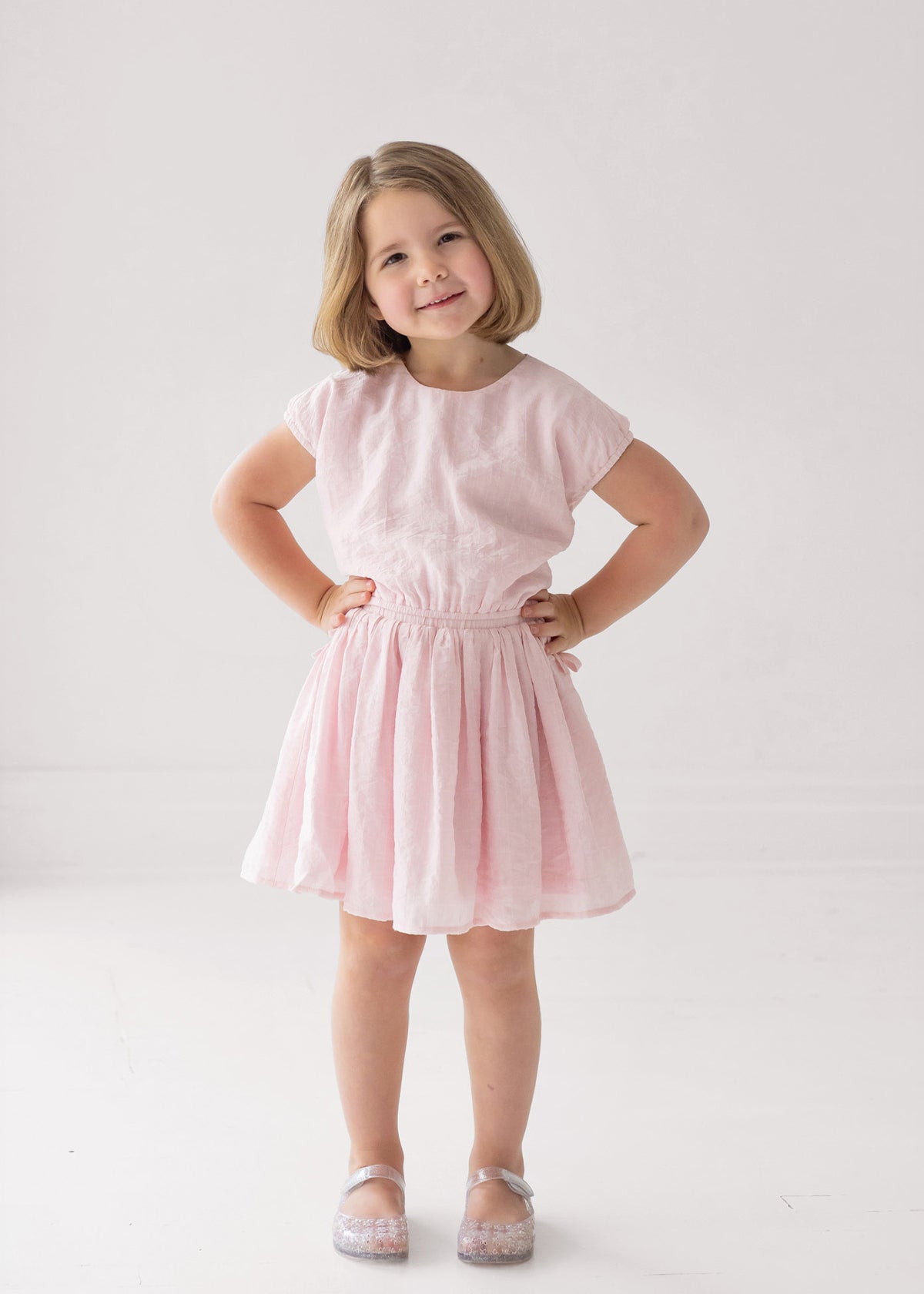 A young girl with short blonde hair smiles, hands on hips, wearing the Mabel and Honey Girlpop Dropshoulder Two Piece Set Pink and sparkly shoes against a plain white background.