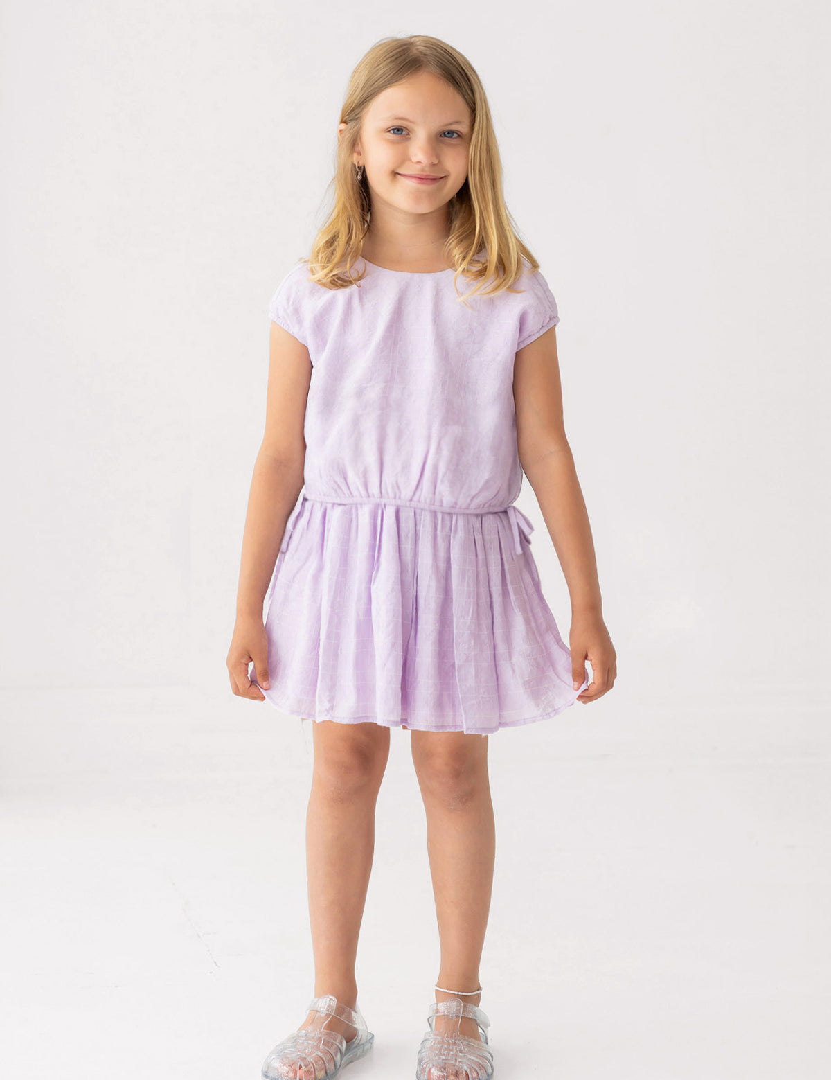 A young blonde girl smiles in a Mabel and Honey Girlypop Two Piece Set Purple with clear jelly sandals, standing against a plain white background.