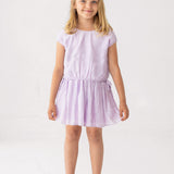 A young blonde girl smiles in a Mabel and Honey Girlypop Two Piece Set Purple with clear jelly sandals, standing against a plain white background.