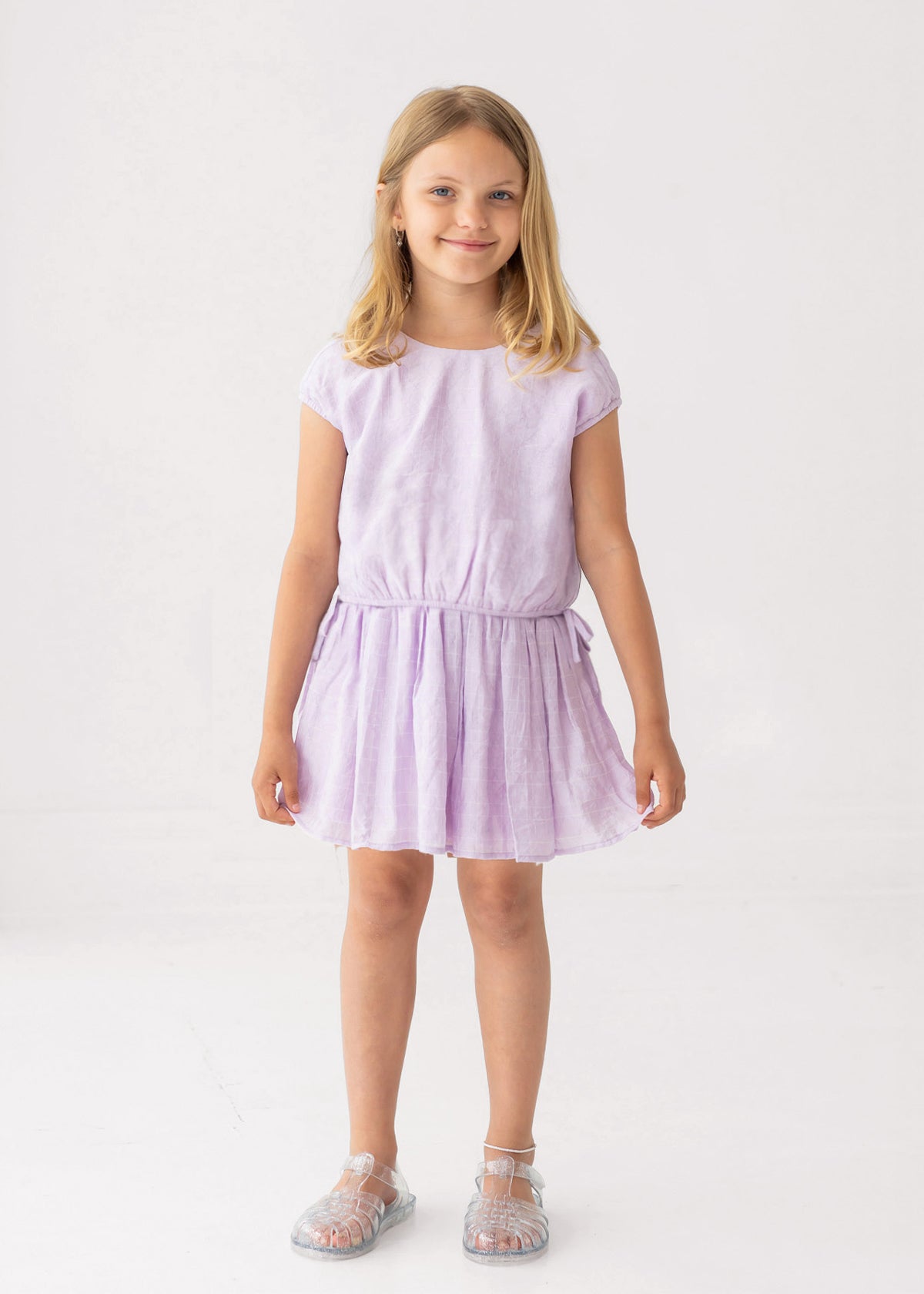 A young blonde girl smiles in a Mabel and Honey Girlypop Two Piece Set Purple with clear jelly sandals, standing against a plain white background.