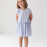 A young girl with short brown hair stands barefoot on a white floor, hands behind her back, slightly smiling and gazing aside, wearing the Mabel and Honey Girlypop Two Piece Set Blue with a cap-sleeve top and matching skirt.
