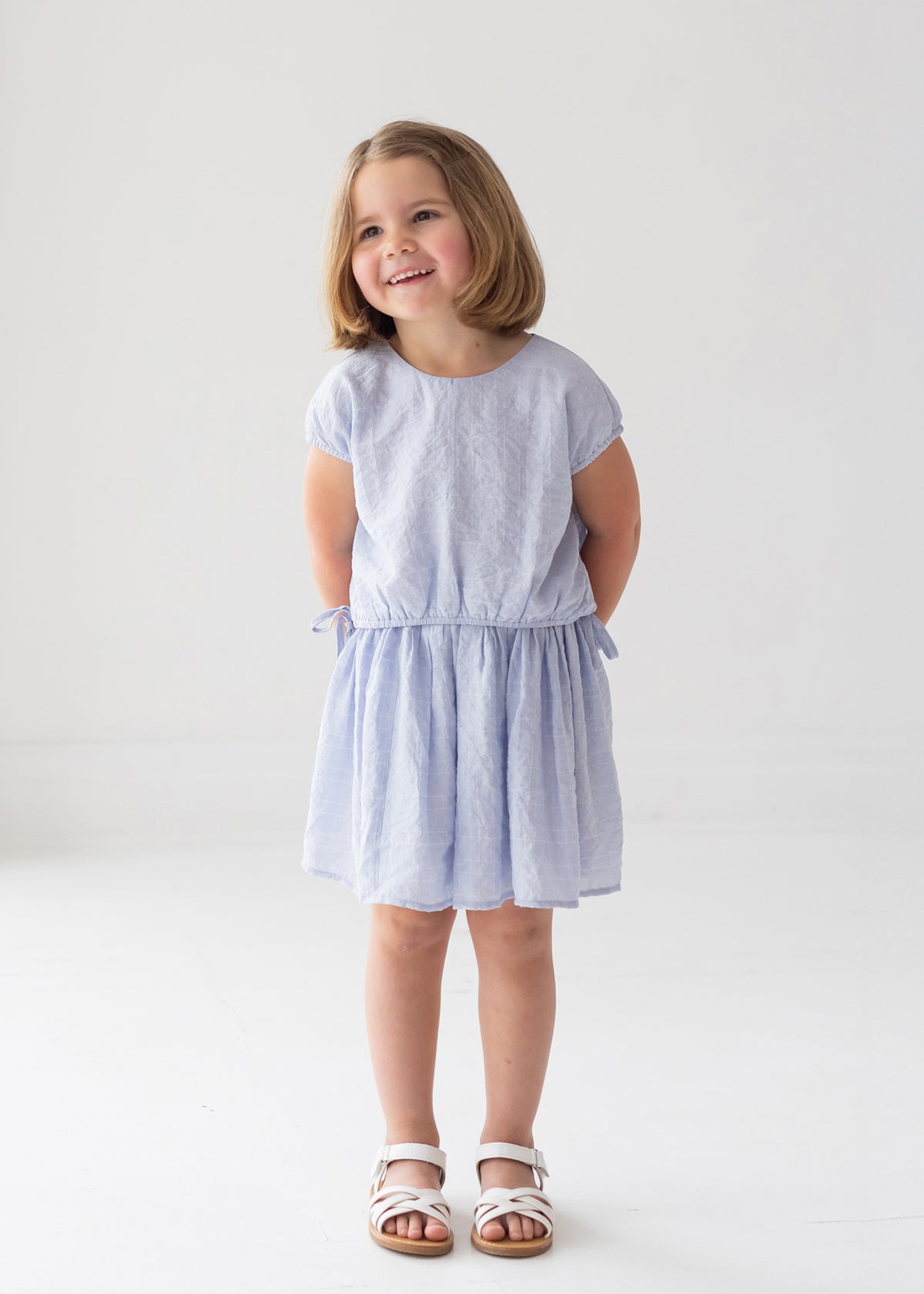 A young girl with short brown hair stands barefoot on a white floor, hands behind her back, slightly smiling and gazing aside, wearing the Mabel and Honey Girlypop Two Piece Set Blue with a cap-sleeve top and matching skirt.