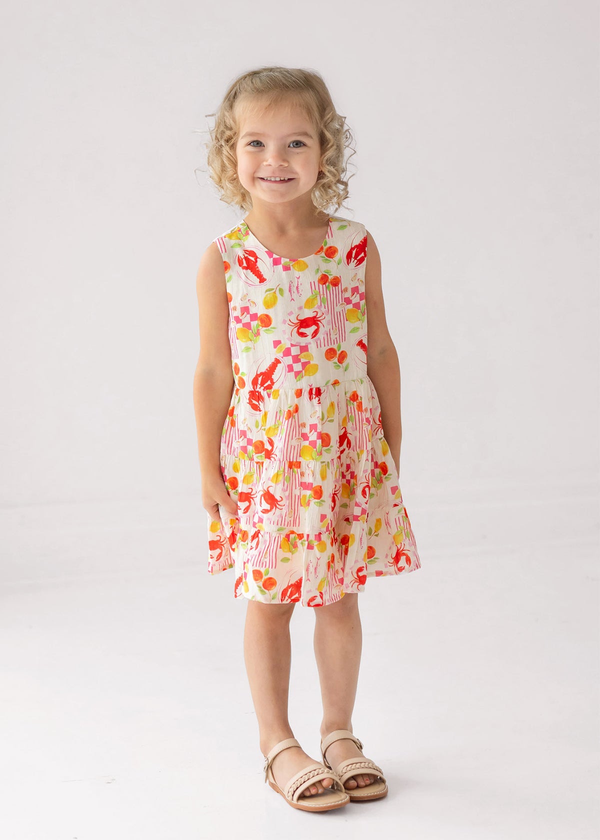 A young girl with curly blonde hair smiles in the Mabel and Honey Coastal Catch Sleeveless Dress, which features a colorful floral and lobster print, paired with light sandals against a plain white background.