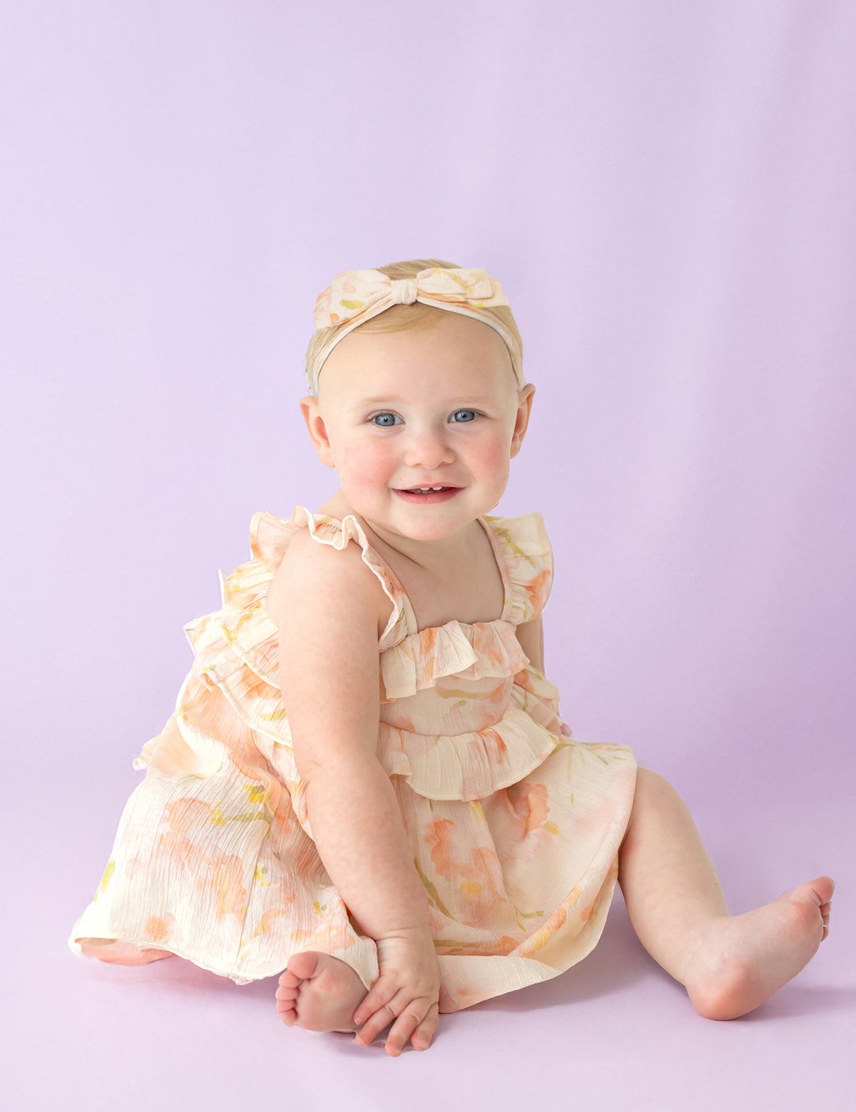 A smiling baby wearing the Isobella and Chloe Sundrop Floral Empire Waist Dress with ruffled straps and a matching headband sits on a light purple background.