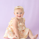 A smiling baby wearing the Isobella and Chloe Sundrop Floral Empire Waist Dress with ruffled straps and a matching headband sits on a light purple background.