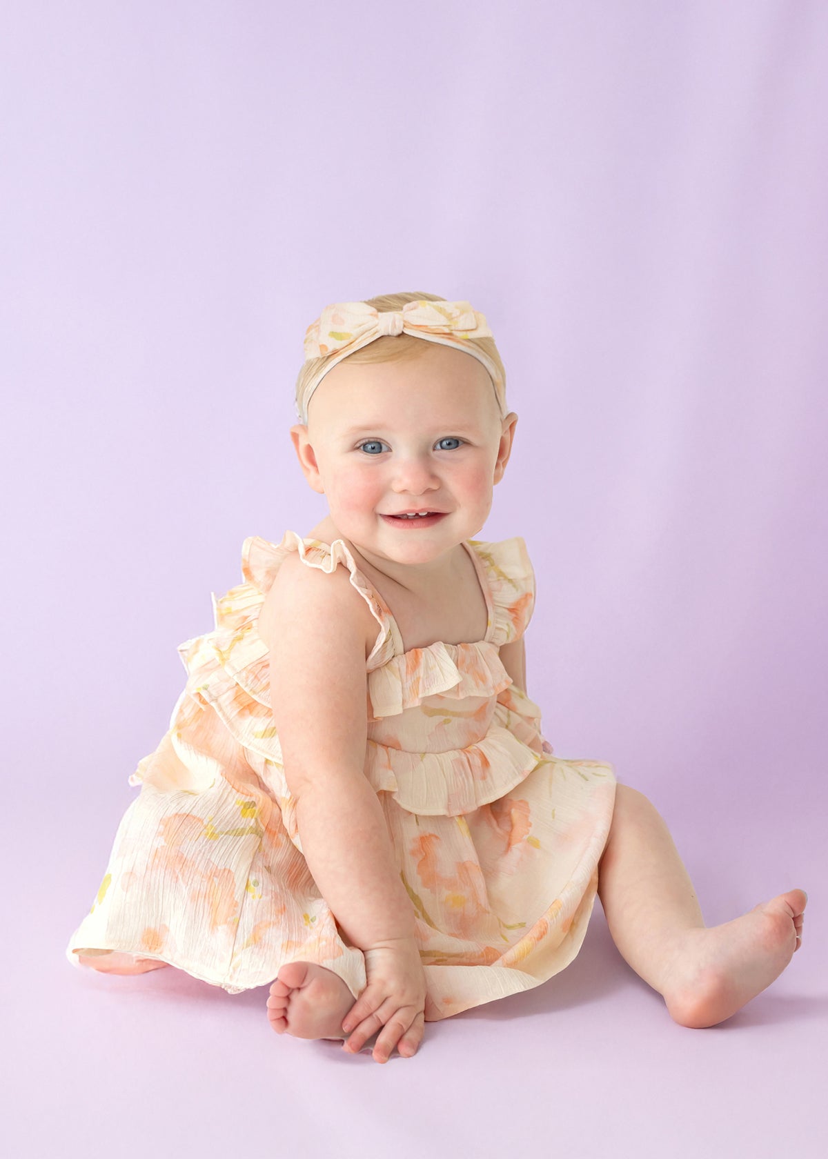 A smiling baby wearing the Isobella and Chloe Sundrop Floral Empire Waist Dress with ruffled straps and a matching headband sits on a light purple background.