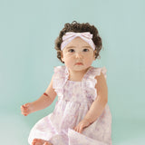A baby with curly dark hair wears the Isobella and Chloe Lilac Dreams Baby Toddler Ruffles Dress with a matching headband, sitting barefoot on a light blue background and looking at the camera.