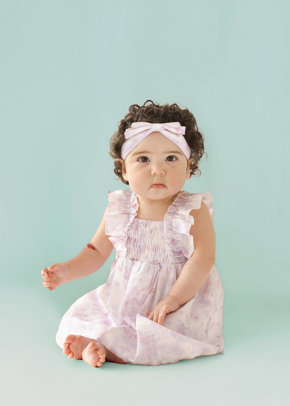 A baby with curly dark hair wears the Isobella and Chloe Lilac Dreams Baby Toddler Ruffles Dress with a matching headband, sitting barefoot on a light blue background and looking at the camera.