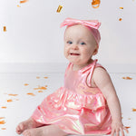 A smiling baby wears the Strawberry Sparkle Sleeveless Baby Dress by Isobella and Chloe with a matching headband, sitting on a white floor surrounded by gold confetti and streamers.