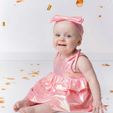 A smiling baby wears the Strawberry Sparkle Sleeveless Baby Dress by Isobella and Chloe with a matching headband, sitting on a white floor surrounded by gold confetti and streamers.