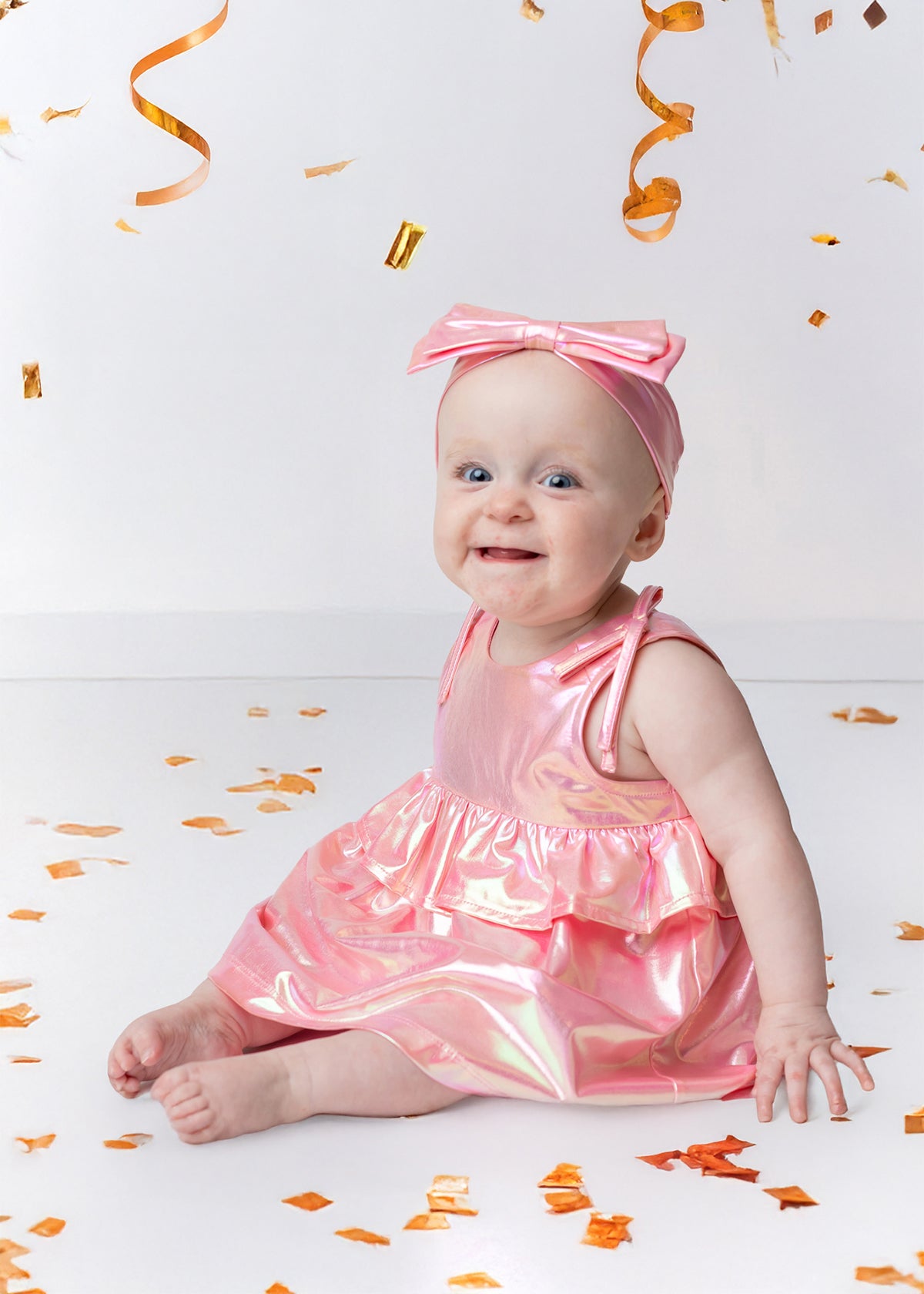 A smiling baby wears the Strawberry Sparkle Sleeveless Baby Dress by Isobella and Chloe with a matching headband, sitting on a white floor surrounded by gold confetti and streamers.