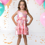 A young girl in the Isobella and Chloe Strawberry Sparkle Sleeveless Dress Pink stands smiling with her hands on her hips, surrounded by teal and pink balloons and gold confetti for a festive atmosphere.
