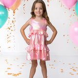 A young girl in the Isobella and Chloe Strawberry Sparkle Sleeveless Dress Pink stands smiling with her hands on her hips, surrounded by teal and pink balloons and gold confetti for a festive atmosphere.