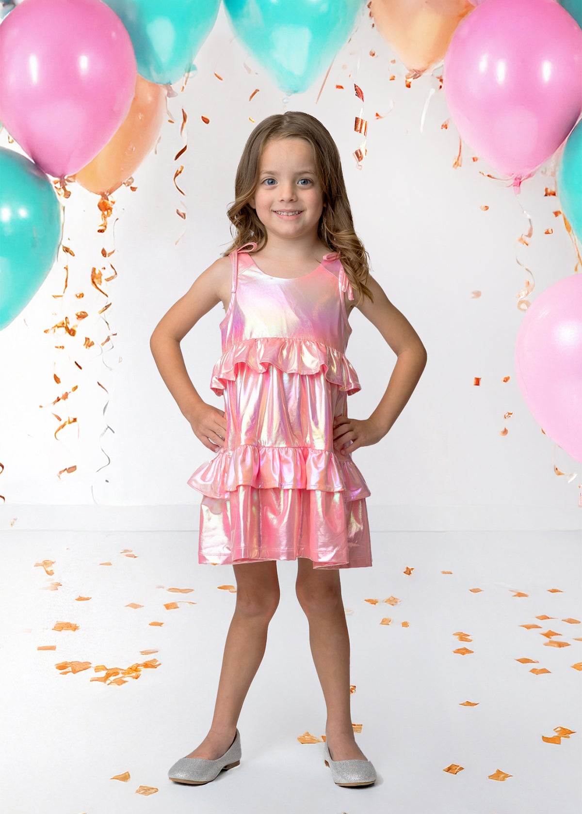A young girl in the Isobella and Chloe Strawberry Sparkle Sleeveless Dress Pink stands smiling with her hands on her hips, surrounded by teal and pink balloons and gold confetti for a festive atmosphere.