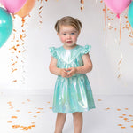 A young child wears the Isobella and Chloe Aquamarine Cap Sleeve Dress and sparkly shoes, standing on a white floor with colorful balloons, gold confetti, and gold streamers hanging above.