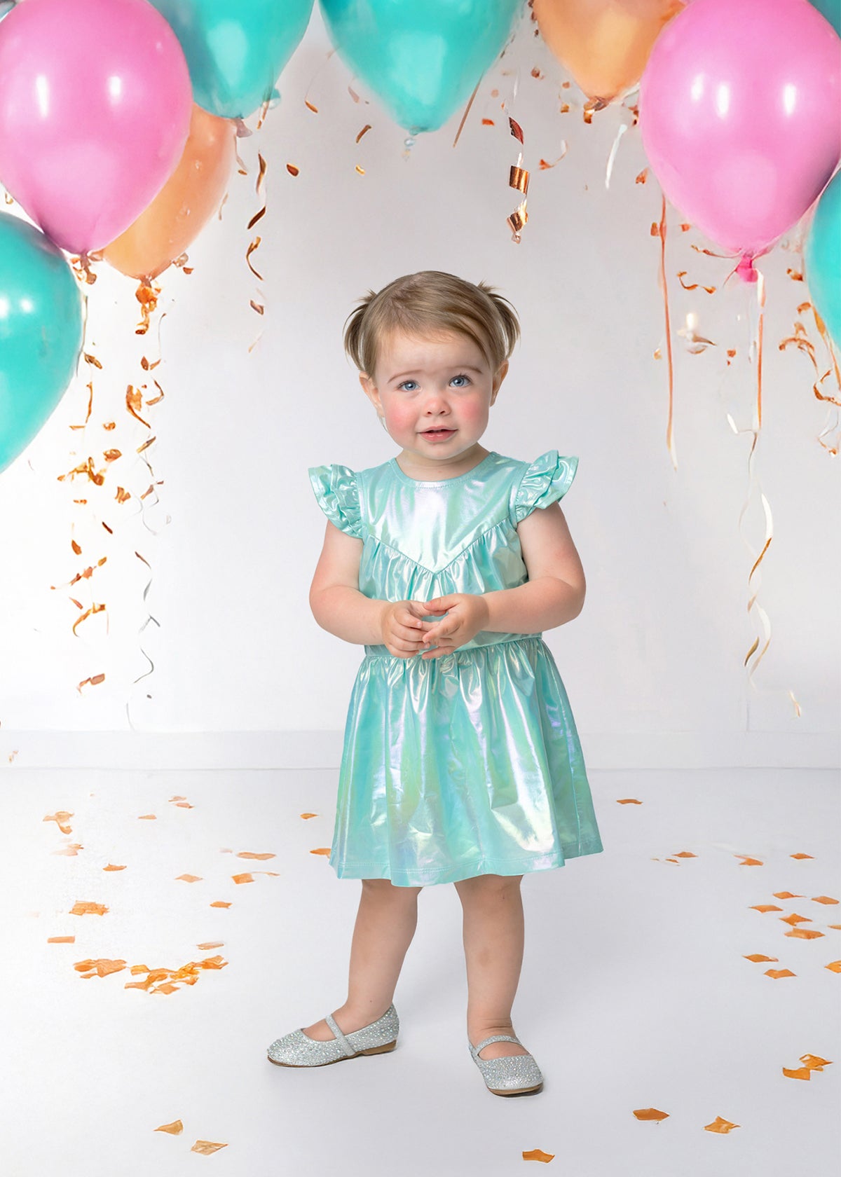 A young child wears the Isobella and Chloe Aquamarine Cap Sleeve Dress and sparkly shoes, standing on a white floor with colorful balloons, gold confetti, and gold streamers hanging above.