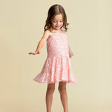 A young girl in the Isobella and Chloe Dazzle Me Blush Sleeveless Dress with gray shoes stands against a beige background, looking down and gently holding out one arm while lifting the hem of her sparkly pink party dress.