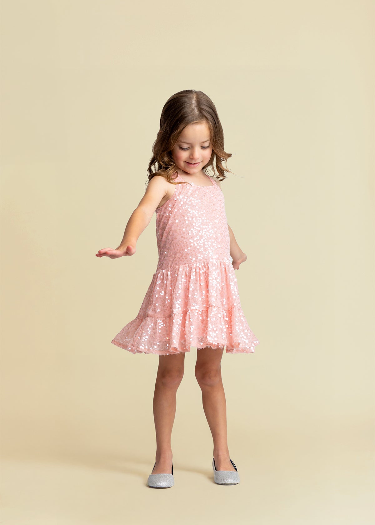 A young girl in the Isobella and Chloe Dazzle Me Blush Sleeveless Dress with gray shoes stands against a beige background, looking down and gently holding out one arm while lifting the hem of her sparkly pink party dress.