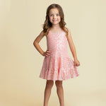 A young girl with long brown hair smiles with one hand on her hip, wearing the Isobella and Chloe Dazzle Me Blush Sleeveless Dress and sparkly beige shoes against a plain beige background.
