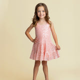 A young girl with long brown hair smiles with one hand on her hip, wearing the Isobella and Chloe Dazzle Me Blush Sleeveless Dress and sparkly beige shoes against a plain beige background.
