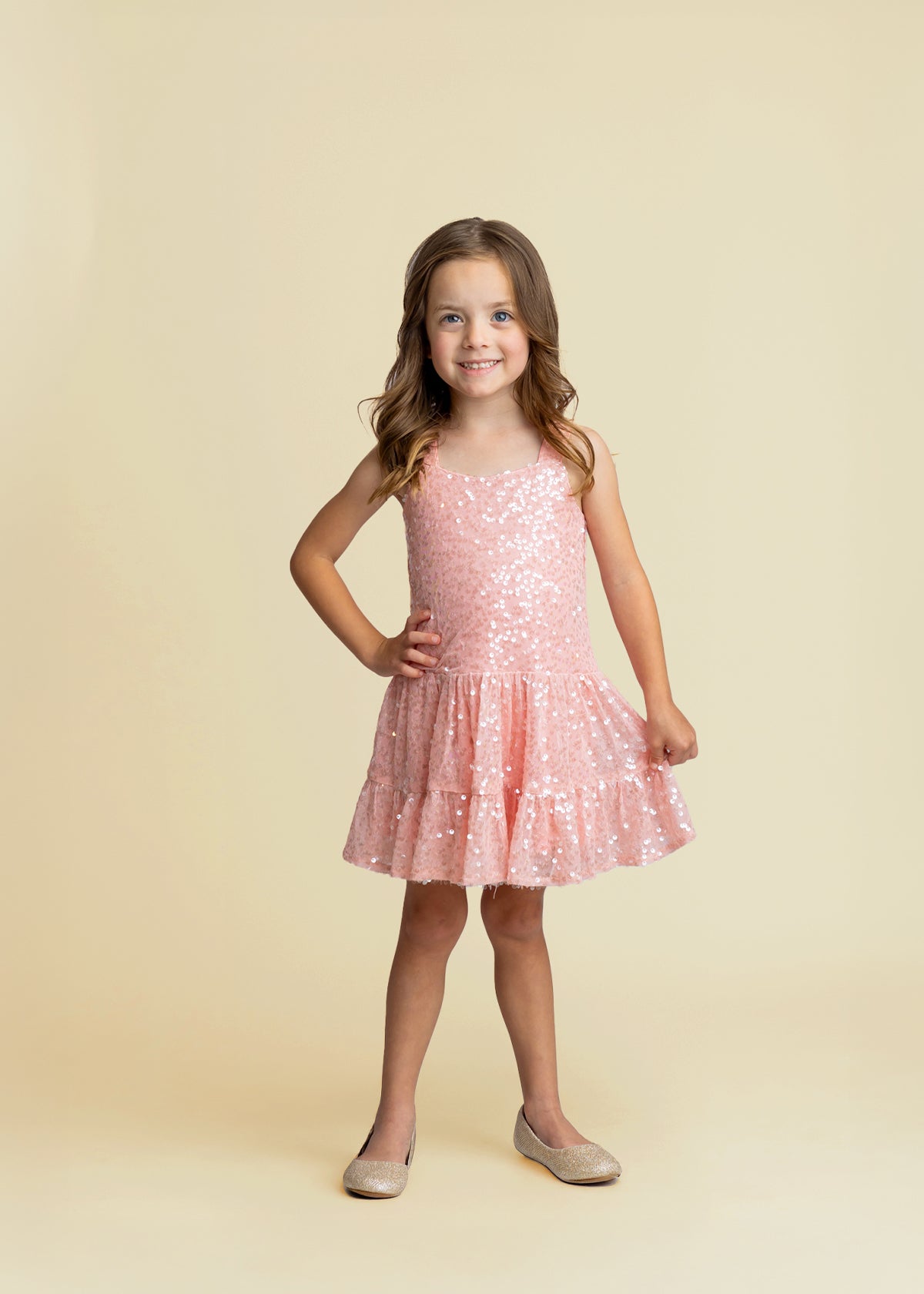 A young girl with long brown hair smiles with one hand on her hip, wearing the Isobella and Chloe Dazzle Me Blush Sleeveless Dress and sparkly beige shoes against a plain beige background.