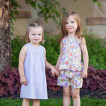 Two young girls stand on the grass, holding hands. One wears a light purple dress and sandals; the other wears the Mabel and Honey Tulip Twirl Tank Two Piece Set. Both smile brightly amid green plants and a tree in the background.