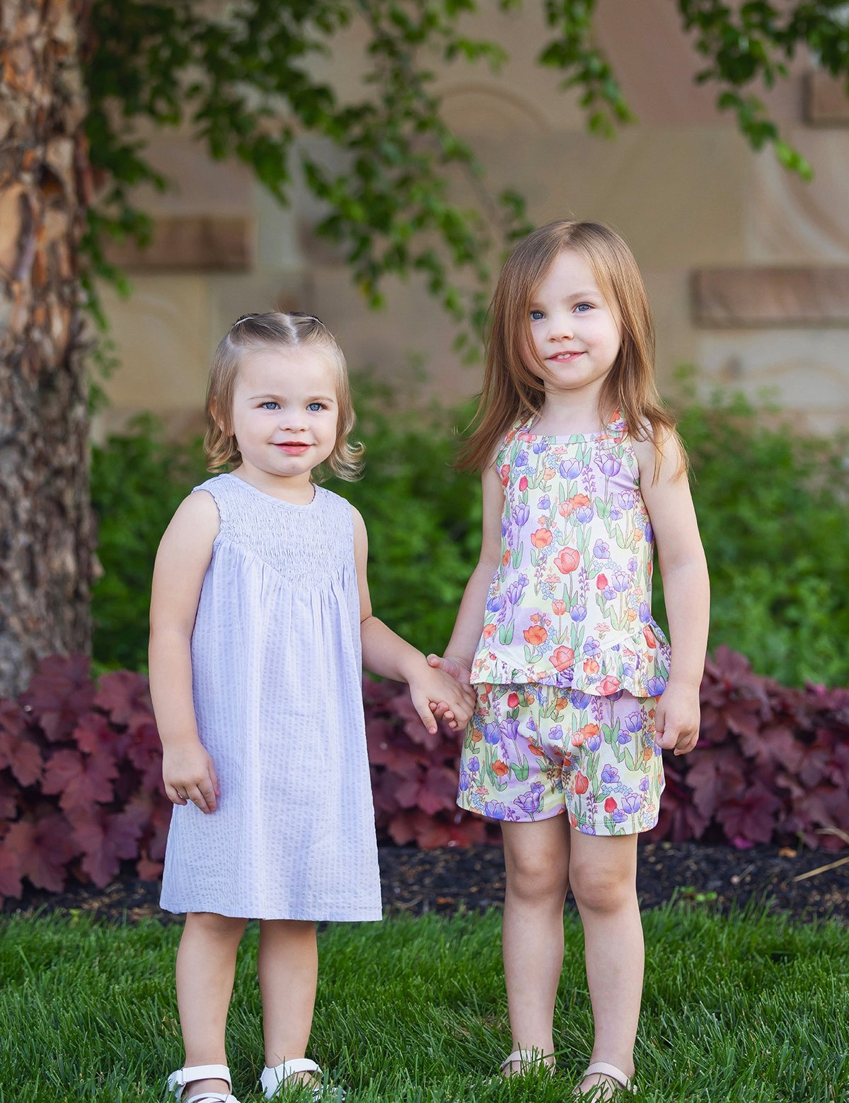 Two young girls stand on the grass, holding hands. One wears a light purple dress and sandals; the other wears the Mabel and Honey Tulip Twirl Tank Two Piece Set. Both smile brightly amid green plants and a tree in the background.