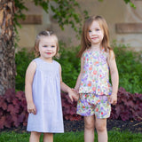 Two young girls stand on the grass, holding hands. One wears a light purple dress and sandals; the other wears the Mabel and Honey Tulip Twirl Tank Two Piece Set. Both smile brightly amid green plants and a tree in the background.
