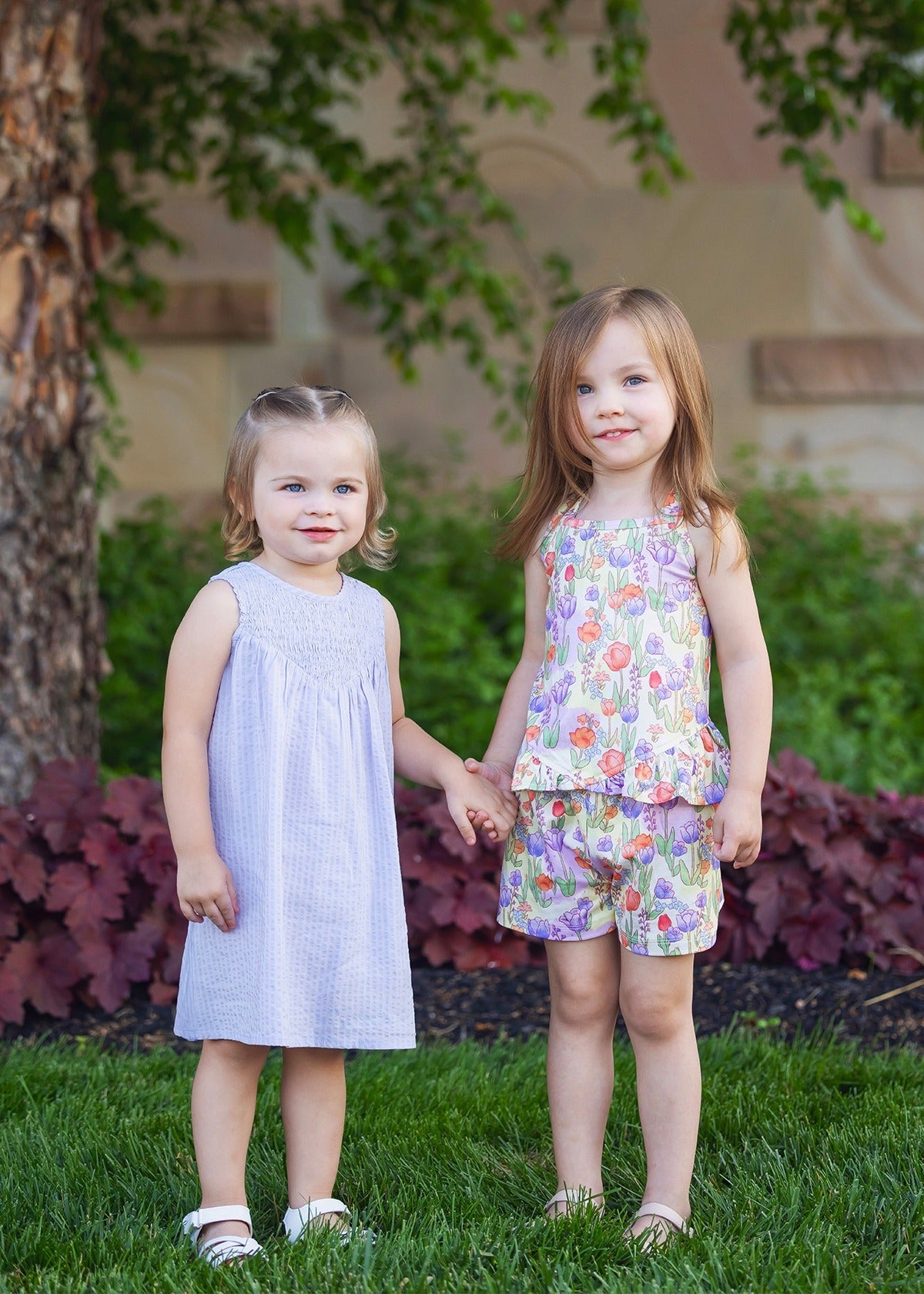 Two young girls stand on the grass, holding hands. One wears a light purple dress and sandals; the other wears the Mabel and Honey Tulip Twirl Tank Two Piece Set. Both smile brightly amid green plants and a tree in the background.