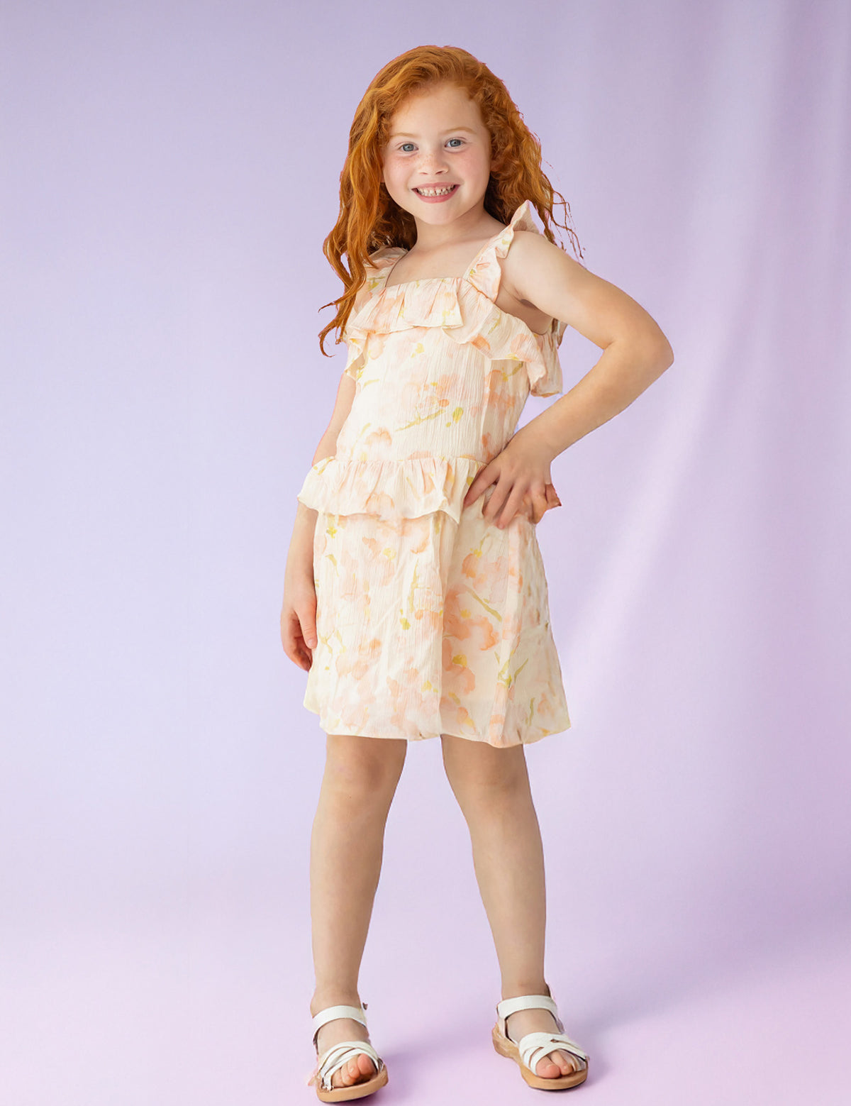 A young girl with long red hair smiles, standing with one hand on her hip in the Isobella and Chloe Sundrop Floral Natural Waist Dress, a sleeveless style with ruffled straps, paired with white sandals against a soft purple background.