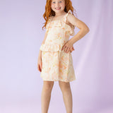 A young girl with long red hair smiles, standing with one hand on her hip in the Isobella and Chloe Sundrop Floral Natural Waist Dress, a sleeveless style with ruffled straps, paired with white sandals against a soft purple background.