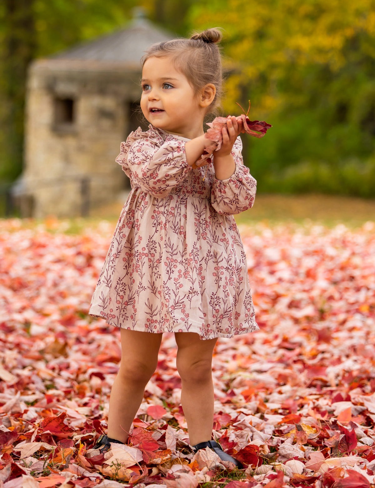 A young girl wears the Mabel + Honey Autumn Floral Tan Long Sleeve Dress, smiling as she stands on red and orange autumn leaves, holding some in her hands with trees and a stone structure in the background.