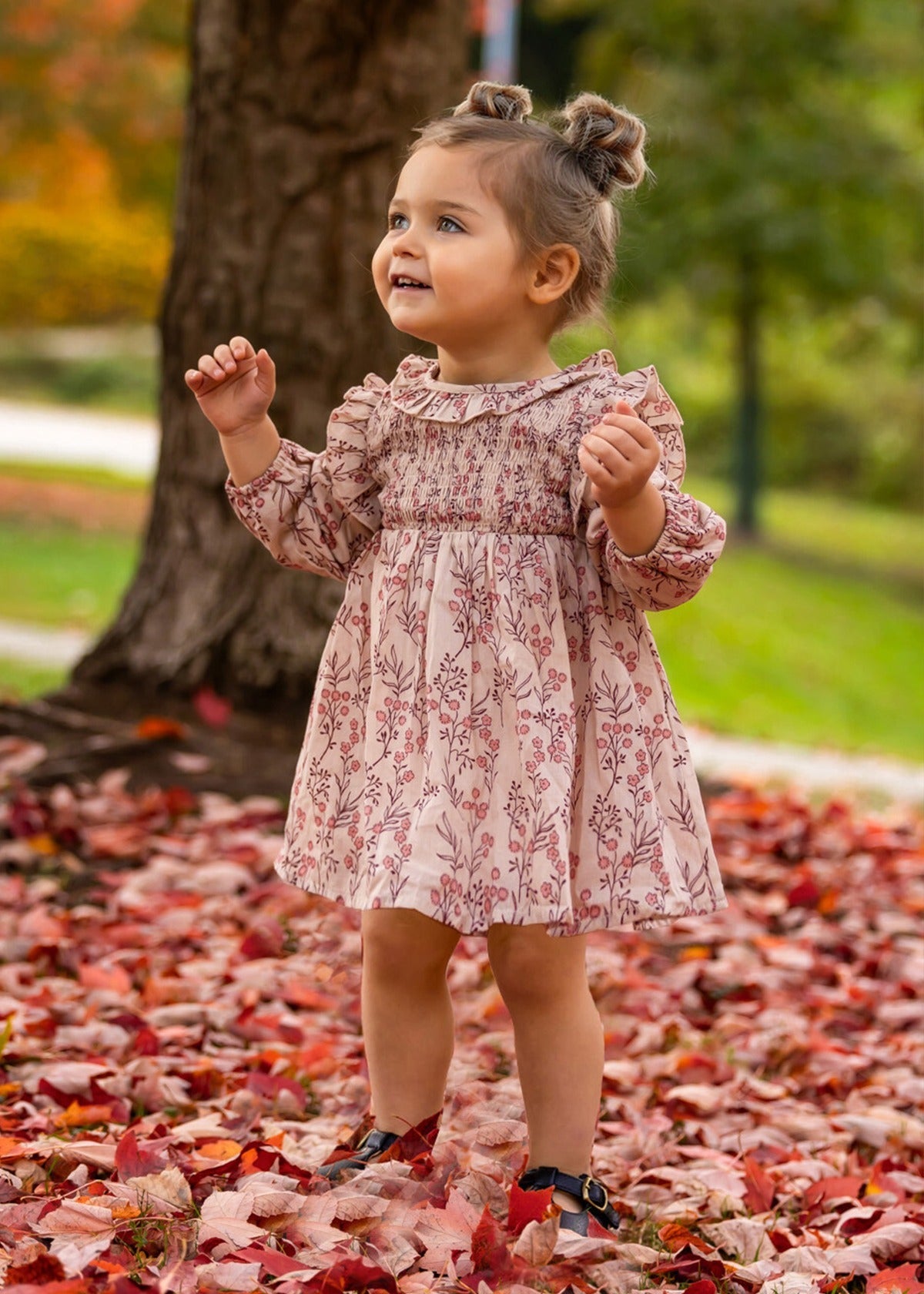 A young girl with brown hair in two buns smiles outdoors among autumn leaves, wearing the Mabel + Honey Autumn Floral Tan Long Sleeve Dress and black shoes. A tree trunk and green foliage are visible in the background.