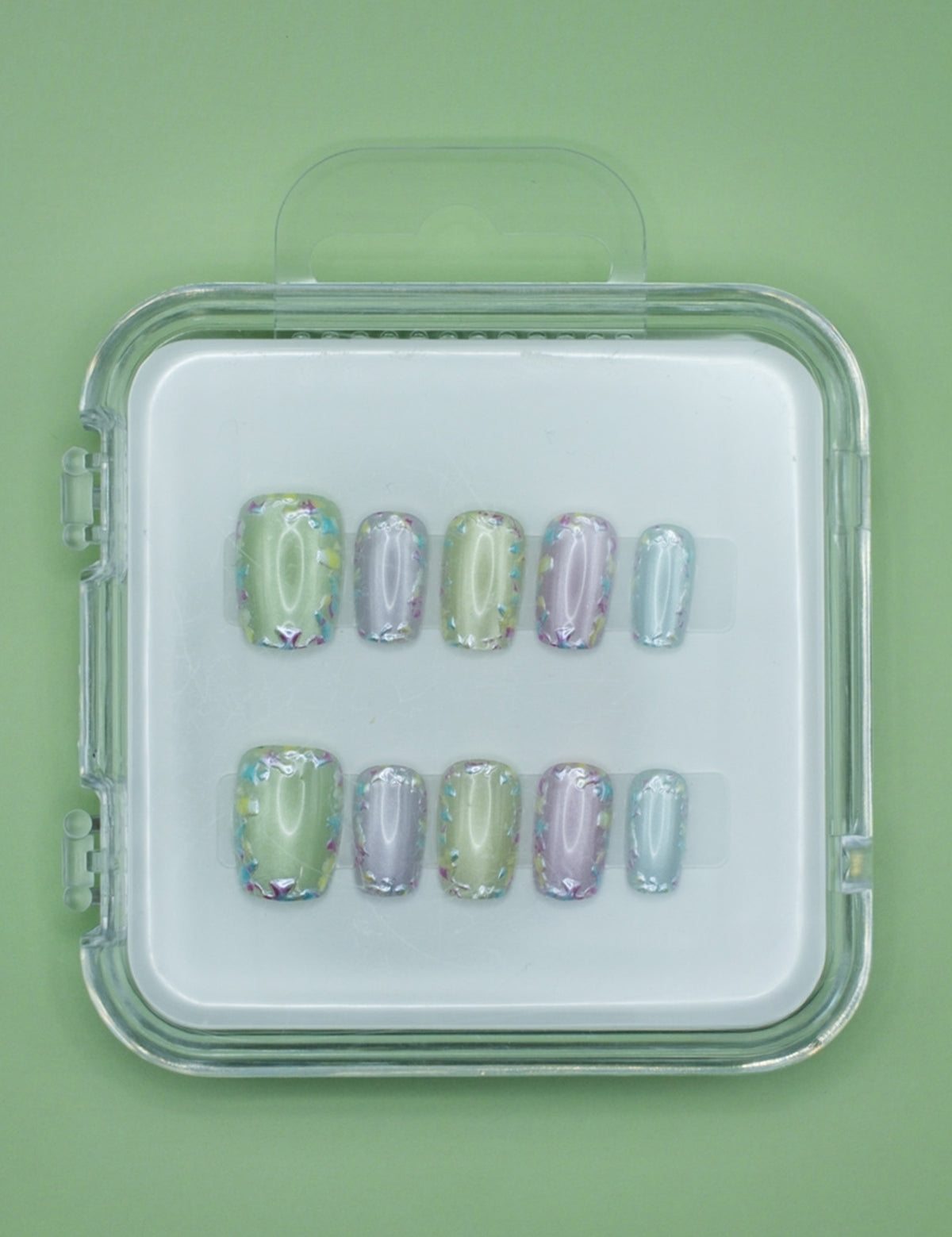 Mabel and Honey’s Confetti set features pastel green, purple, and blue salon-quality press-on nails with iridescent holographic accents, displayed in a clear case against a matching green background.