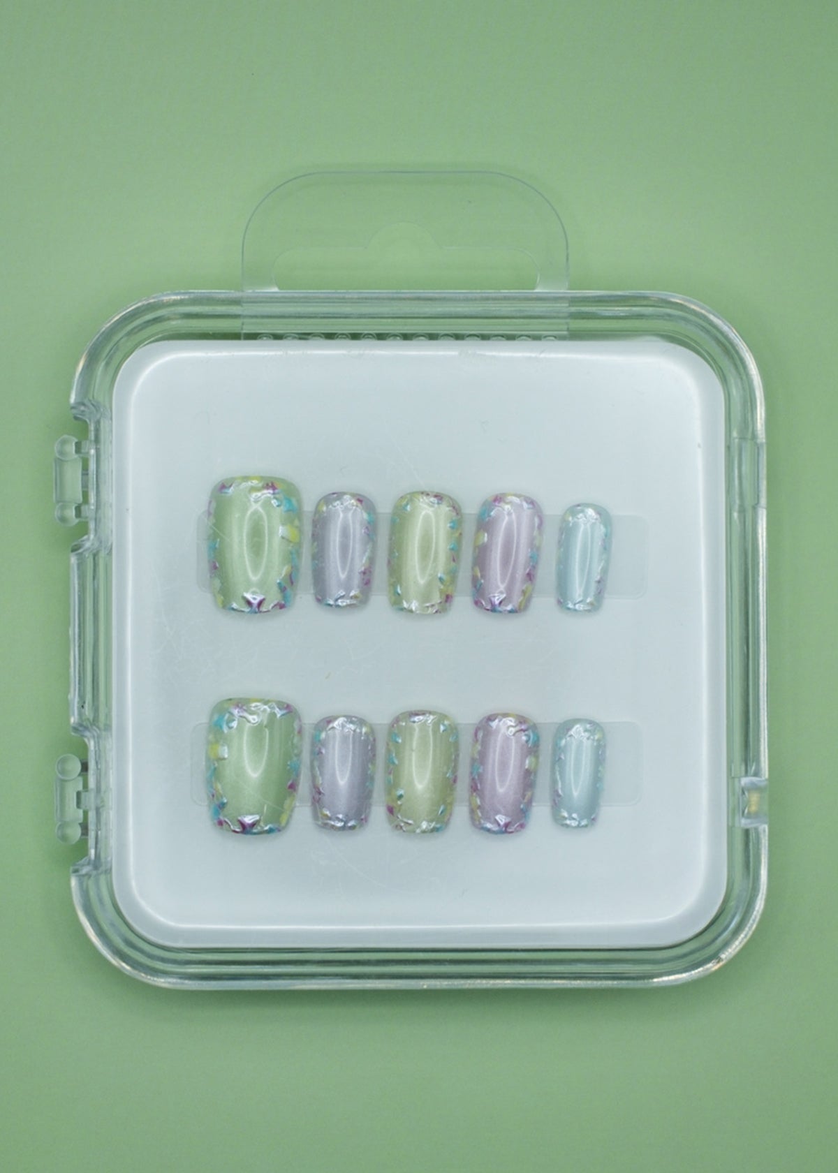 Mabel and Honey’s Confetti set features pastel green, purple, and blue salon-quality press-on nails with iridescent holographic accents, displayed in a clear case against a matching green background.