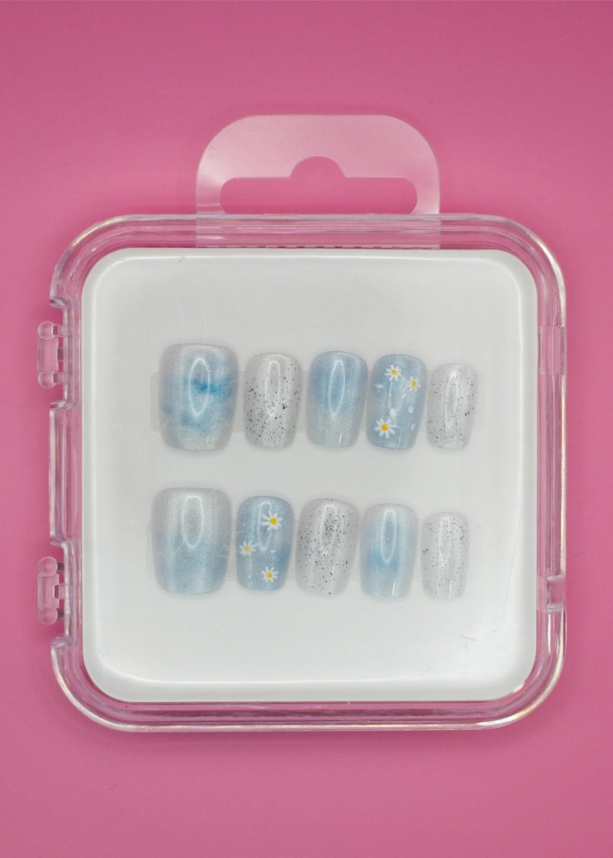 Darling Daisies by Mabel and Honey features blue reusable press-ons with white flower designs and black speckles, shown in a clear plastic case against a pink background.