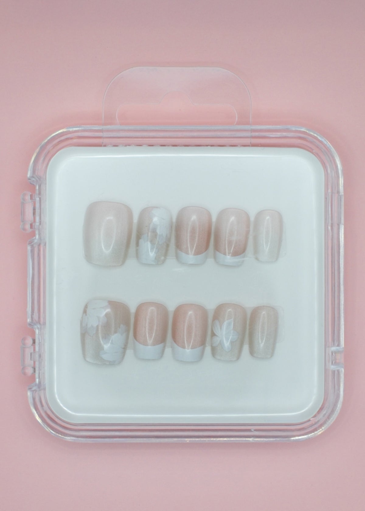 Mabel and Honey's White Floral press-on nails come in a clear case, featuring a pale pink base with white floral details and French tips, displayed on a light pink background.
