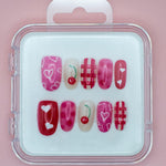 Cherry Cherry Lady by Mabel and Honey is a reusable nail set with pink and red press-ons in a clear case, featuring hearts, cherries, and plaid designs on a light pink background.