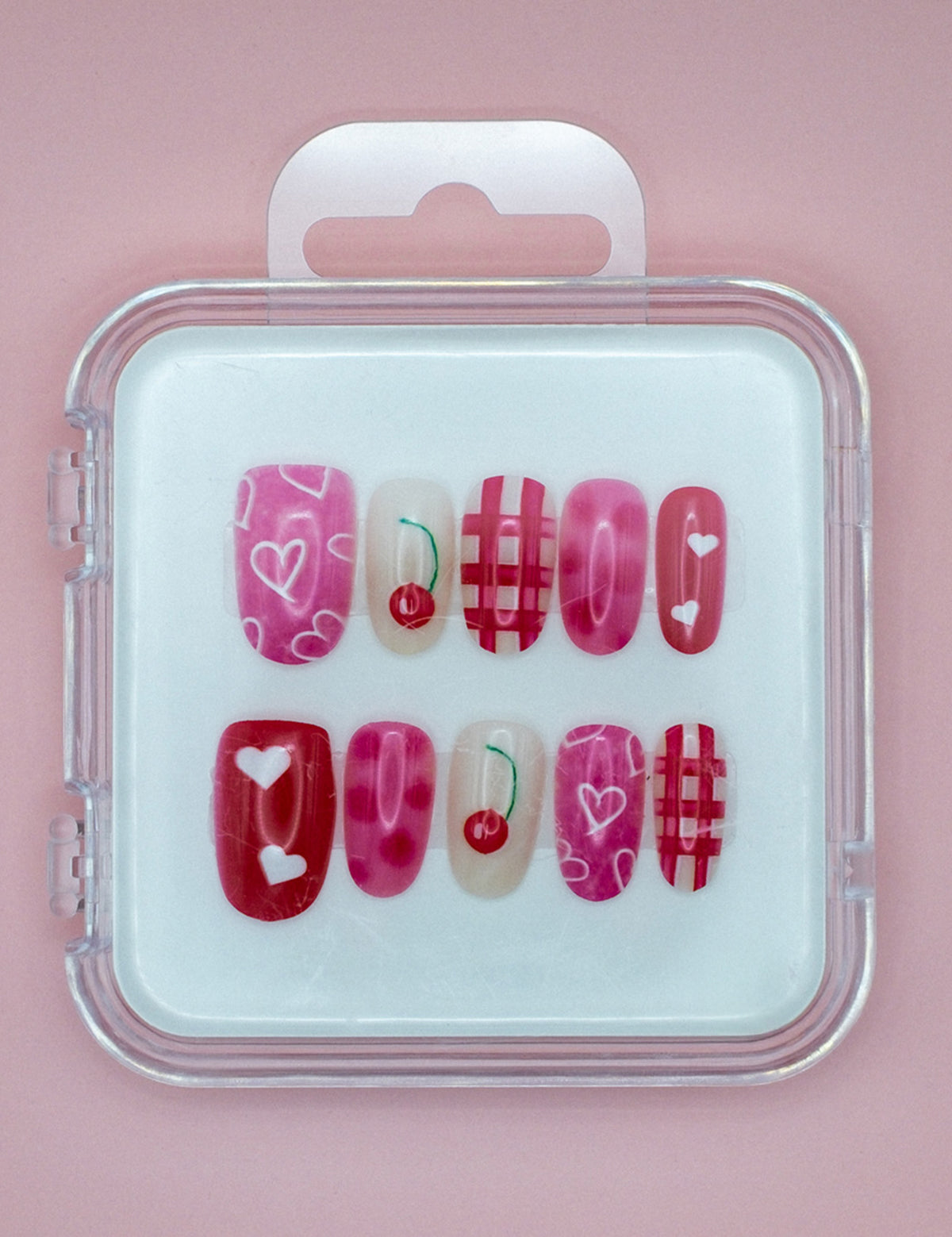 Cherry Cherry Lady by Mabel and Honey is a reusable nail set with pink and red press-ons in a clear case, featuring hearts, cherries, and plaid designs on a light pink background.