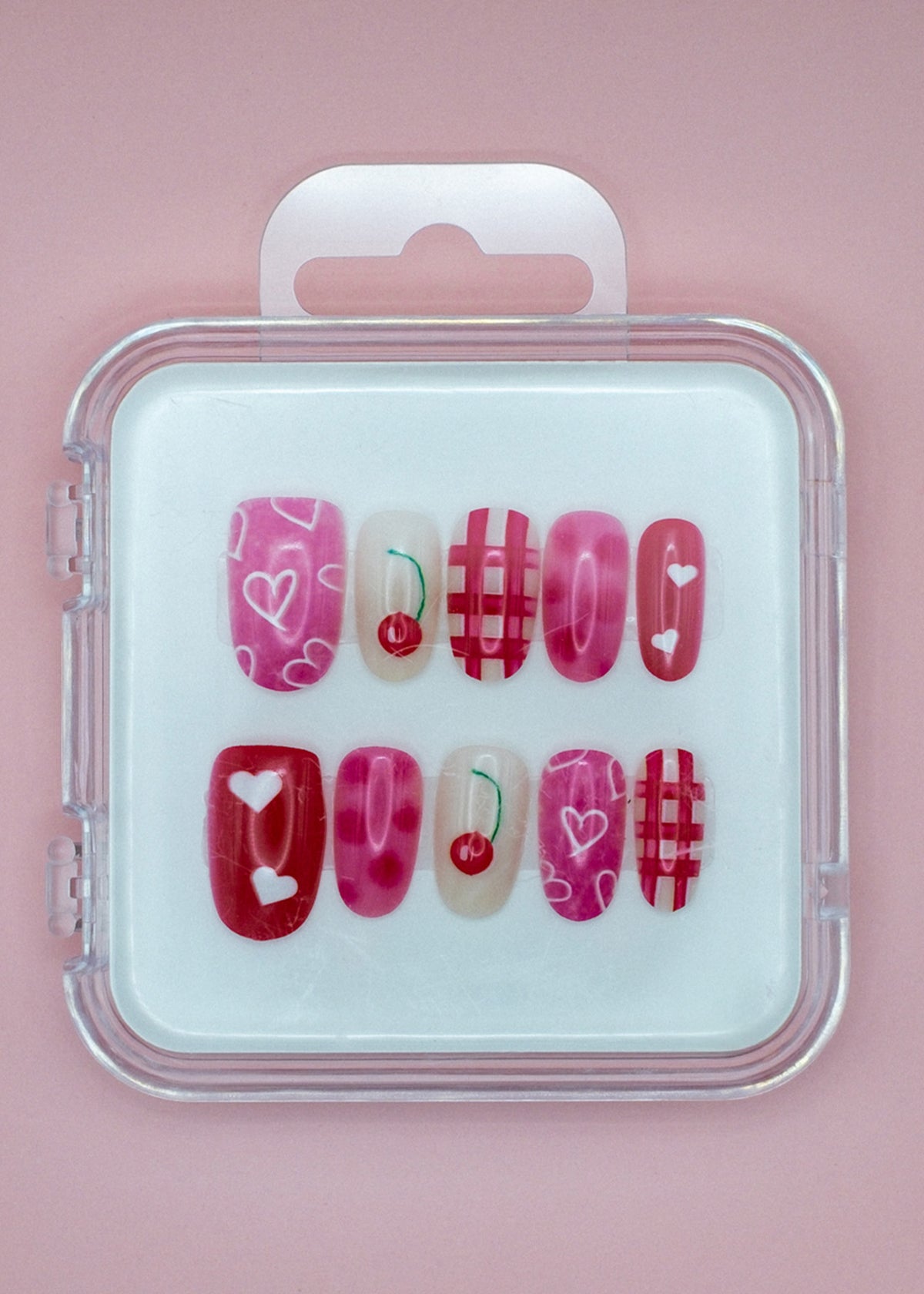 Cherry Cherry Lady by Mabel and Honey is a reusable nail set with pink and red press-ons in a clear case, featuring hearts, cherries, and plaid designs on a light pink background.