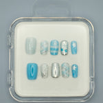 Winter Wonderland by Mabel and Honey features reusable press-on nails in a clear case with silver, blue, and white snowflake, stripe, plaid, and solid designs—perfect for salon-quality manicures at home. Arranged neatly on a gray background.