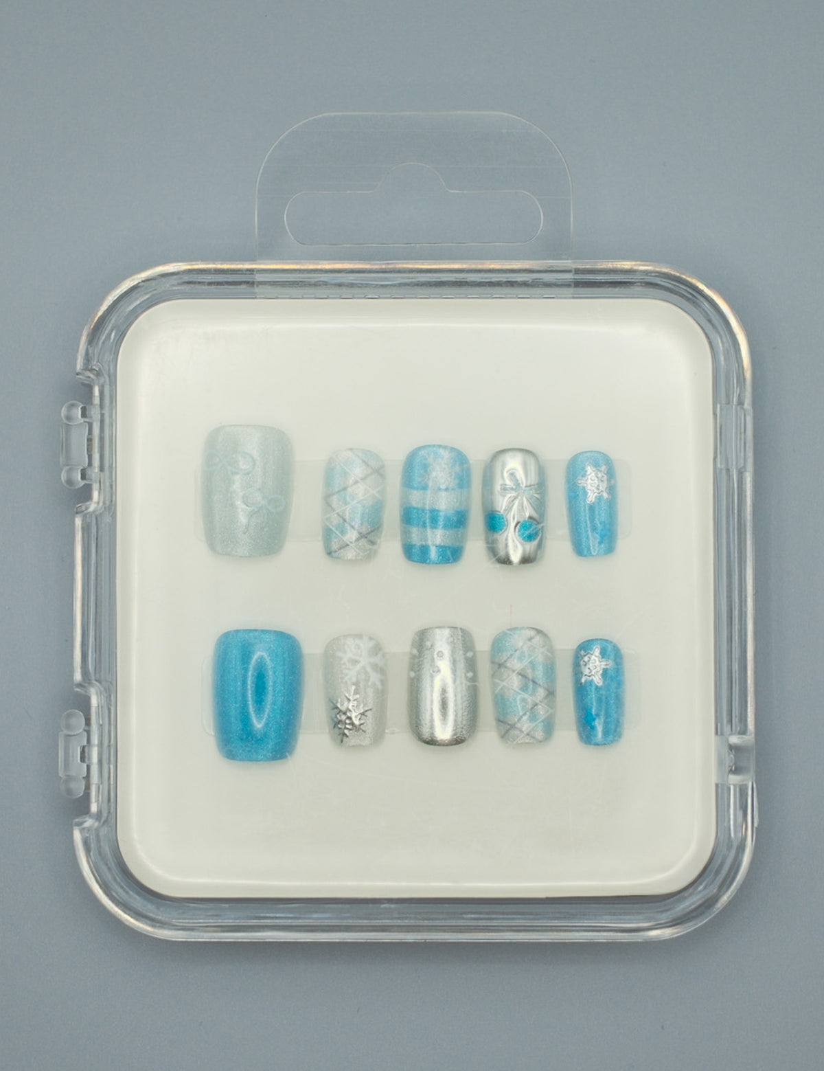 Winter Wonderland by Mabel and Honey features reusable press-on nails in a clear case with silver, blue, and white snowflake, stripe, plaid, and solid designs—perfect for salon-quality manicures at home. Arranged neatly on a gray background.