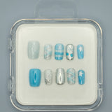 Winter Wonderland by Mabel and Honey features reusable press-on nails in a clear case with silver, blue, and white snowflake, stripe, plaid, and solid designs—perfect for salon-quality manicures at home. Arranged neatly on a gray background.